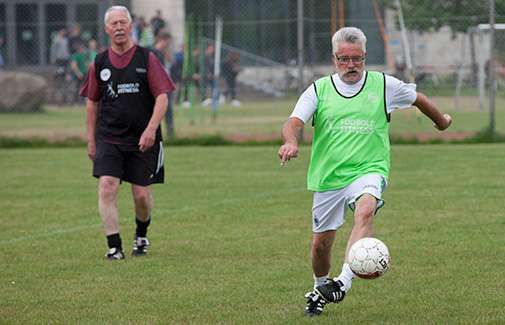 Två äldre män spelar fotboll på en gräsplan, varav en dribblar bollen.